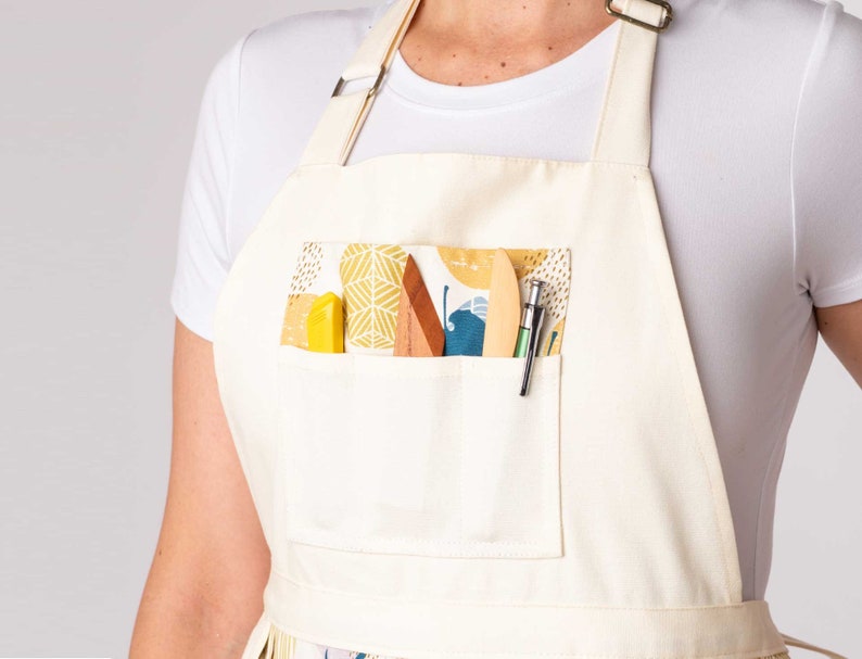 K&ouml;nnte beinhalten: Eine wei&szlig;e Leinensch&uuml;rze mit einer gro&szlig;en Tasche auf der Vorderseite. Die Tasche ist mit einem farbenfrohen gemusterten Stoff gef&uuml;ttert und enth&auml;lt verschiedene Werkzeuge, darunter ein Lineal aus Holz, ein Metallschaber und ein Bleistift.
