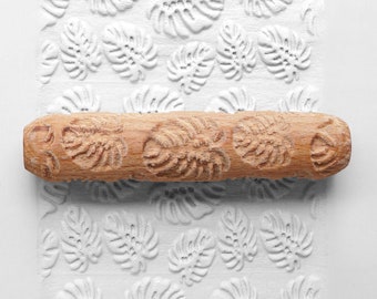 May include: A wooden rolling pin with a carved leaf pattern. The rolling pin is used to create an embossed leaf design on a white surface.