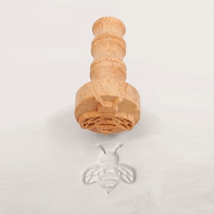 Può includere: Un timbro di legno a forma di ape con un design dettagliato sulla testa del timbro e un'impressione di ape scolpita. Il timbro ha un manico cilindrico ed è realizzato in legno chiaro. L'impressione dell'ape è visibile su una superficie bianca.