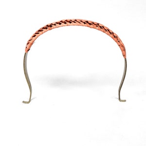 Może przedstawiać: Brzoskwiniowa pleciona opaska do włosów z metalową ramą. Pleciona część tworzy łuk na górze, a metalowa rama rozciąga się w dół z zakrzywionym kształtem i małymi, płaskimi końcami. Opaska do włosów jest na białym tle.