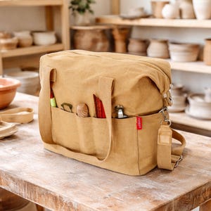 May include: A tan canvas tote bag with multiple external pockets, a shoulder strap, and top handles. The pockets hold various tools, including a red-handled knife and a wooden spoon. The bag has a zipped top and a small red tag with white text.