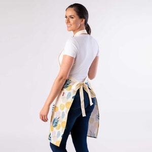 K&ouml;nnte beinhalten: Eine Frau tr&auml;gt ein wei&szlig;es T-Shirt und eine blaue Jeans und tr&auml;gt eine cremefarbene Sch&uuml;rze mit einem bunten abstrakten Muster. Die Sch&uuml;rze hat einen Bindeg&uuml;rtel in der Taille.