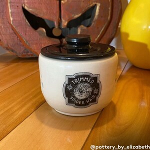 May include: A white ceramic jar with a black lid and a black and white label that reads "Trimmed Spider Hair".