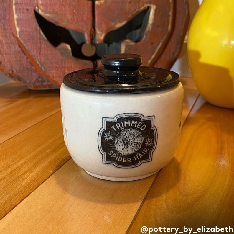 May include: A white ceramic jar with a black lid and a black and white label that reads "Trimmed Spider Hair".