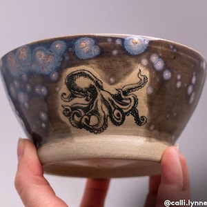 Op de afbeelding: Een keramische kom met een donkerbruine glazuur en witte stippen. De kom heeft een zwart-witte illustratie van een octopus.