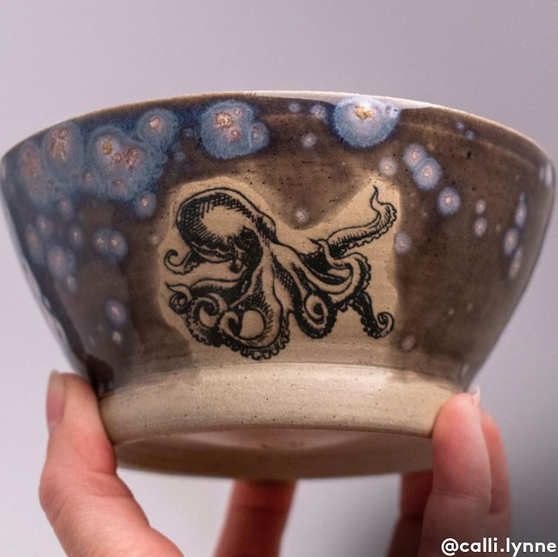 Op de afbeelding: Een keramische kom met een donkerbruine glazuur en witte stippen. De kom heeft een zwart-witte illustratie van een octopus.
