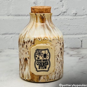 May include: A ceramic jar with a cork lid and a label that reads "Black Widow Cobweb Stew". The jar is decorated with a brown and white glaze and has a spider web design on the label.