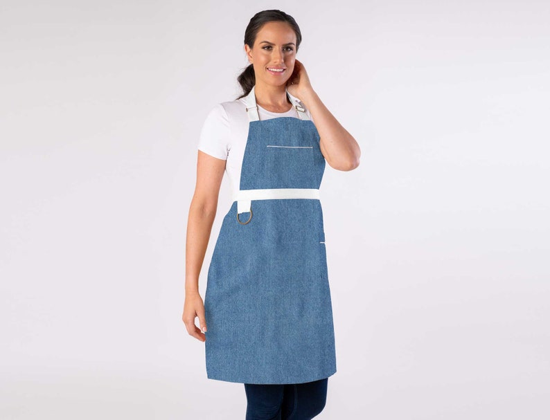 May include: A blue denim apron with white straps and a white pocket. The apron has a metal ring on the strap.