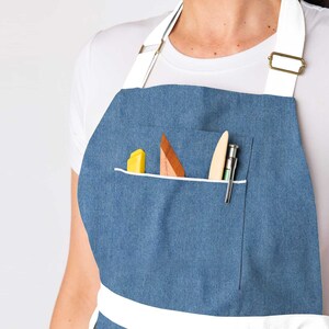 May include: A blue denim apron with a white trim and a large pocket. The pocket contains a yellow highlighter, a wooden tool, and a pen.