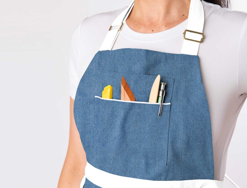 May include: A blue denim apron with a white trim and a large pocket. The pocket contains a yellow highlighter, a wooden tool, and a pen.