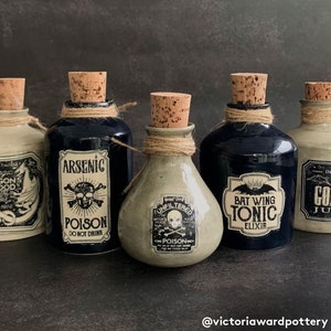May include: Five small ceramic bottles with cork stoppers and twine tied around the necks. Each bottle has a different label with a spooky Halloween theme. The labels include text such as "Arsenic", "Poison", "Bat Wing Tonic", "Unfiltered", and "Goblin Juice".
