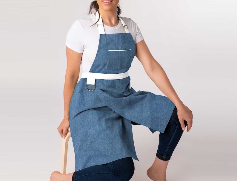May include: A blue denim apron with white straps and a white pocket. The apron has a brass ring on the strap.
