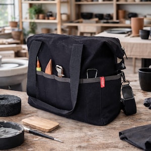 Puede incluir: Una bolsa de herramientas de lona negra con múltiples bolsillos, una correa para el hombro y asas. La bolsa contiene varias herramientas de cerámica, incluyendo una espátula de madera, una herramienta de metal y un lápiz. La palabra "SANBAD" está impresa en una etiqueta roja en la bolsa.