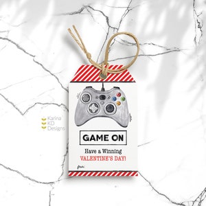 Puede incluir: Etiqueta de regalo a rayas rojas y blancas con un dibujo de un mando de videojuegos gris. La etiqueta dice "Game On" y "Have a Winning Valentine's Day!"