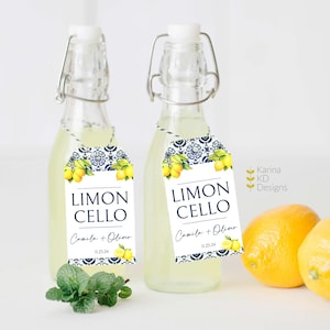 May include: Two small glass bottles filled with a clear liquid, each with a white tag that reads "Limon Cello" in black text. The tags also feature a watercolor illustration of lemons and blue and white patterned tiles. The text "Camila + Oliver" and the date "11.23.24" are printed on the tags.