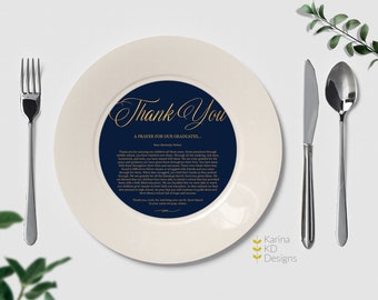 May include: A white ceramic plate with a navy blue center featuring gold script that reads "Thank You" and a prayer for graduates. Silverware, including a fork, knife, and spoon, is arranged around the plate.