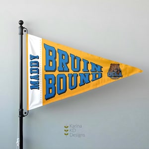 May include: A yellow pennant with the words "BRUIN BOUND" in blue block letters. The pennant also features the UCLA Bruins logo and the name "MADDY" in blue. The pennant is mounted on a black pole.