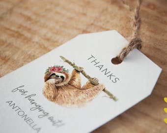 May include: A white gift tag with a watercolor illustration of a sloth wearing a floral crown and hanging from a branch. The tag reads "THANKS for hanging out! ANTONELLA" in a handwritten font, with a jute string attached.