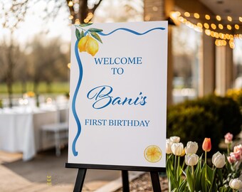 May include: A white welcome sign on a black easel, with the text "WELCOME TO Bani's FIRST BIRTHDAY" in blue script. The sign is decorated with a lemon and blue ribbon design. The sign is outdoors with flowers in the foreground.