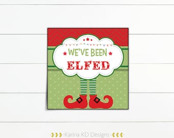 Op de afbeelding: Een rood en groen printbaar bord met de tekst "We've Been Elfed" in een witte wolk met rode sterren en een lichtsnoer. Het bord heeft een paar elfenbenen en rode schoenen.