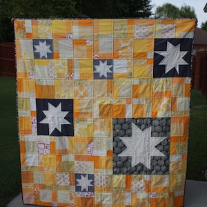 May include: A patchwork quilt in a yellow, orange and white colour scheme. The quilt features a variety of star designs in white on a black background.