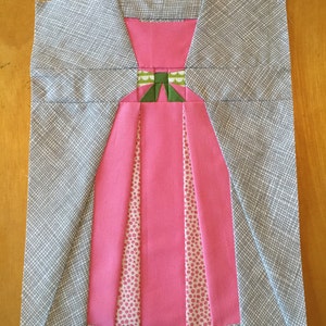May include: A pink dress block with a green bow and white polka dots on the skirt. The dress block is on a gray background.