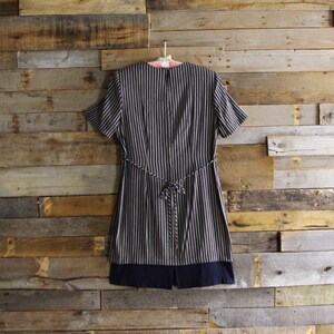 May include: A short-sleeved dress with vertical black and white stripes, a matching tie belt, and a solid navy blue hem. The dress is hanging on a pink hanger against a rustic wooden background.