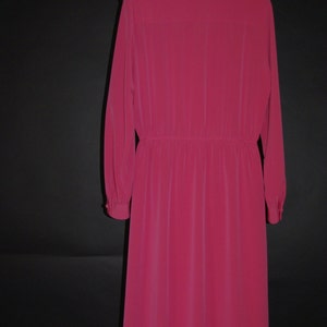 May include: A long-sleeved, knee-length dress in a vibrant fuchsia color. The dress features a buttoned cuff, a gathered waist, and a classic collar. The dress is hanging on a black metal hanger against a black background.
