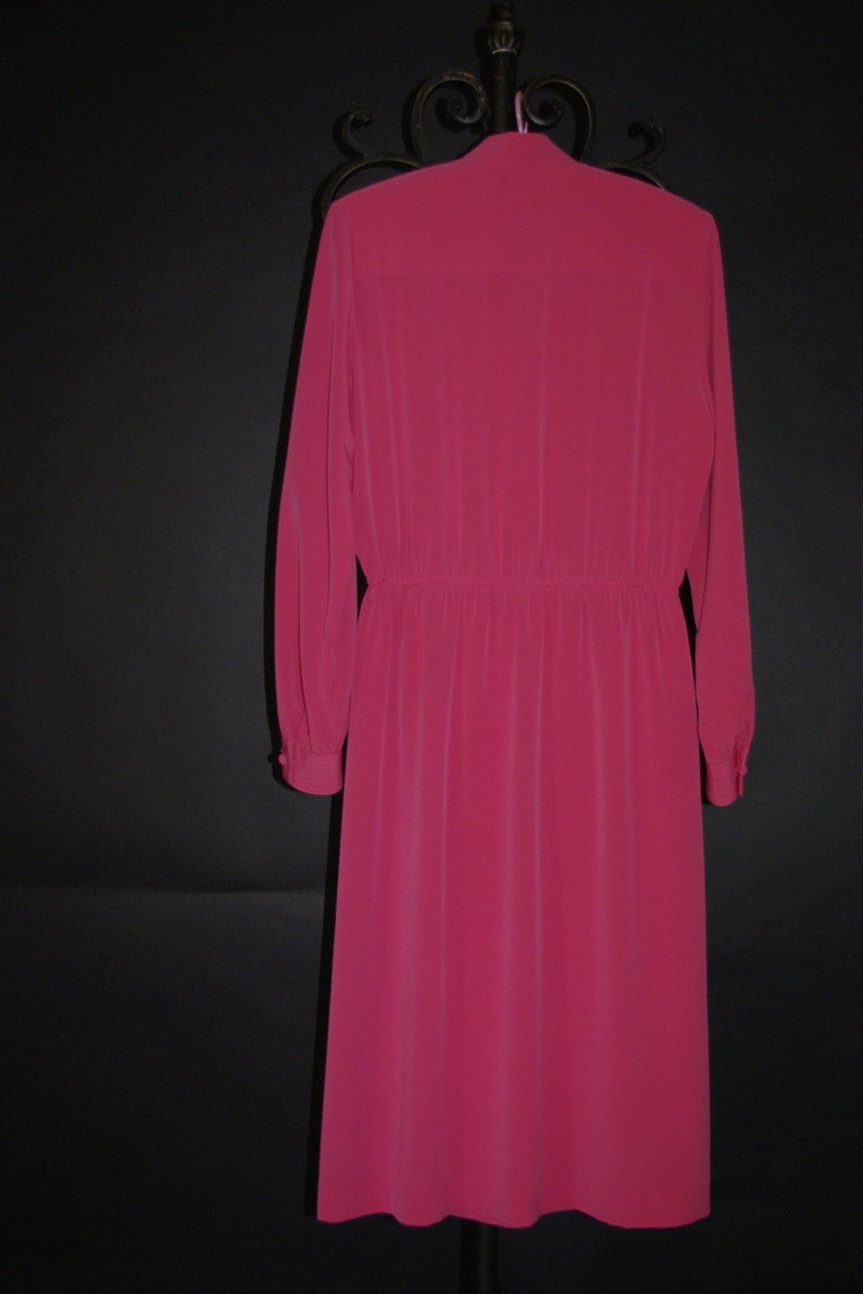 May include: A long-sleeved, knee-length dress in a vibrant fuchsia color. The dress features a buttoned cuff, a gathered waist, and a classic collar. The dress is hanging on a black metal hanger against a black background.
