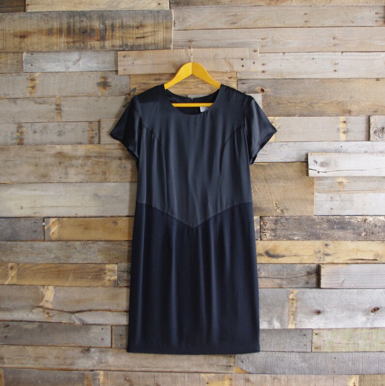May include: A dark navy blue dress with short sleeves hangs on a yellow hanger against a wooden plank background. The dress features a contrasting panel design at the waist, creating a visual V-shape.