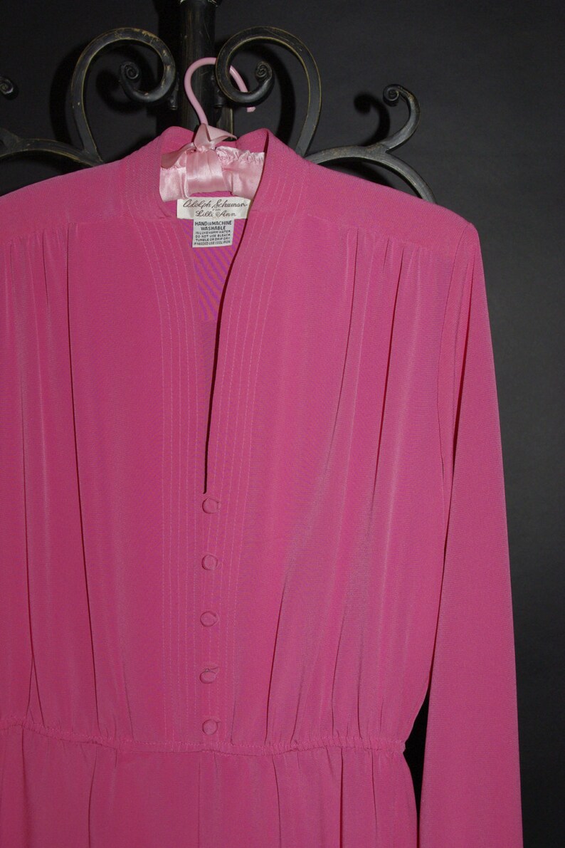 May include: A bright pink dress with long sleeves and a mandarin collar, hanging on a pink hanger. The dress features vertical pleats down the front and a row of buttons. The label reads "Adolph Schuman Lilli Ann".