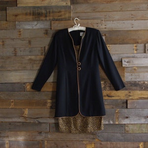 May include: A black long-sleeve dress with a contrasting tan trim and two gold buttons. The dress has a leopard print pattern at the bottom. The dress is hanging on a white hanger against a wooden plank background.
