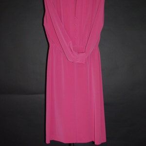 May include: A vibrant fuchsia dress with long sleeves and a button-down front. The dress features a high neckline and a fitted waist, hanging on a decorative metal hanger. The dress is made of a smooth, flowing fabric, perfect for a formal occasion.