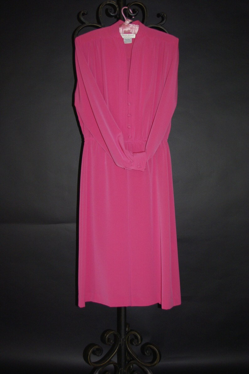 May include: A vibrant fuchsia dress with long sleeves and a button-down front. The dress features a high neckline and a fitted waist, hanging on a decorative metal hanger. The dress is made of a smooth, flowing fabric, perfect for a formal occasion.
