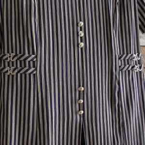 May include: Close-up of a vertically striped garment in navy blue and off-white. The fabric has a textured appearance. The garment features silver-colored buttons and decorative buckles on the pockets. The overall style suggests a vintage or classic aesthetic.