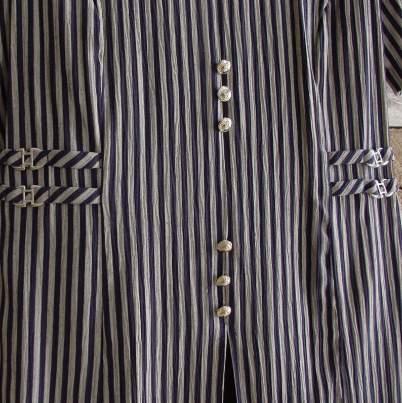 May include: Close-up of a vertically striped garment in navy blue and off-white. The fabric has a textured appearance. The garment features silver-colored buttons and decorative buckles on the pockets. The overall style suggests a vintage or classic aesthetic.