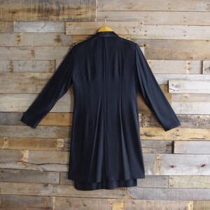 May include: A navy blue long coat with long sleeves hangs on a yellow hanger against a wooden plank background. The coat has a tailored design with a fitted waist and a flared skirt. The back of the coat is visible.