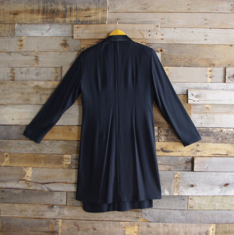 May include: A navy blue long coat with long sleeves hangs on a yellow hanger against a wooden plank background. The coat has a tailored design with a fitted waist and a flared skirt. The back of the coat is visible.