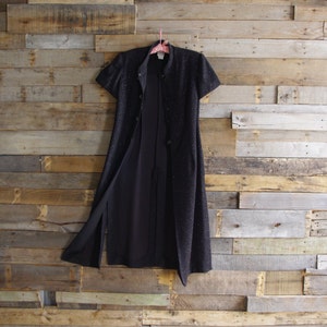 May include: A black dress with short sleeves and a button-up front, hanging on a pink hanger. The dress has a subtle sparkle and a slit up the front. The background is a wooden wall.