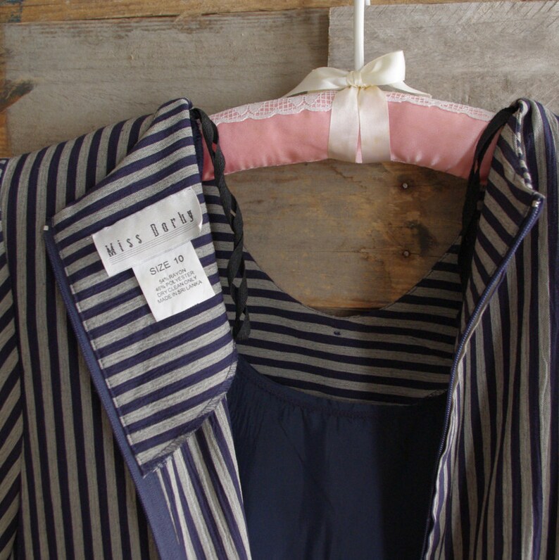 May include: A vintage dress with vertical gray and navy blue stripes, hanging on a pink padded hanger with a white bow. The dress has a label that reads "Miss Darby" and "Size 10". The dress is made of rayon and polyester.