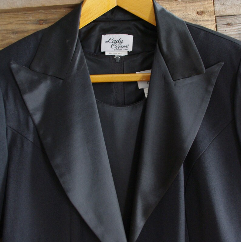 May include: A black two-piece suit with a satin lapel and the label "Lady Carol" visible. The suit is hanging on a wooden hanger against a wooden background. The suit appears to be made of a smooth, dark fabric.