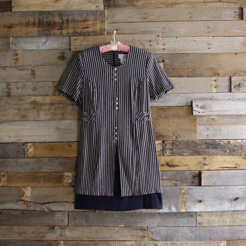 May include: A vintage striped dress with short sleeves, a round neckline, and gold-colored buttons. The dress features vertical stripes in navy and cream, with decorative details at the waist. The hemline is a solid navy blue.