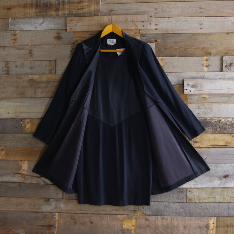 May include: A dark navy blue coat with a classic design, hanging on a wooden hanger. The coat is open, revealing a matching navy blue interior lining. The coat has a tailored fit and a collar.