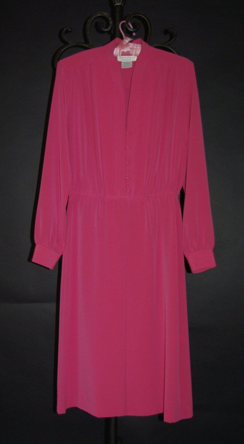May include: A long-sleeved, fuchsia-colored dress with a high collar and button-down front. The dress features a gathered waist and long sleeves with buttoned cuffs. The dress is hanging on a decorative hanger.