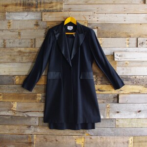 May include: A long, dark navy blazer with a satin lapel and pocket details hangs on a yellow wooden hanger against a rustic wood background. The blazer has a single-button closure and a tailored fit, suitable for formal or business attire.