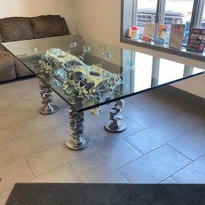May include: A glass-top table with a unique design. The base of the table is made from a car engine crankshaft. The table is in a room with a brown leather couch and a window.