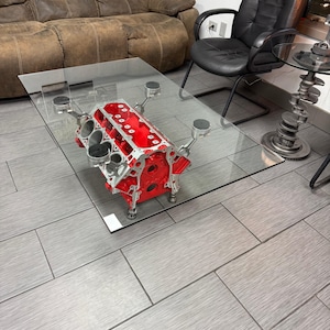 May include: A unique coffee table featuring a red engine block base supporting a clear glass top. The engine block is detailed with silver pistons and components. The table is in a room with a brown sofa and black chair.