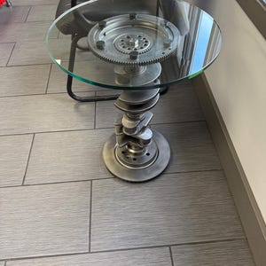 May include: A unique, industrial-style side table. It features a round, clear glass top supported by a base constructed from repurposed automotive parts. The base includes a crankshaft and a gear, creating a distinctive, metallic aesthetic.