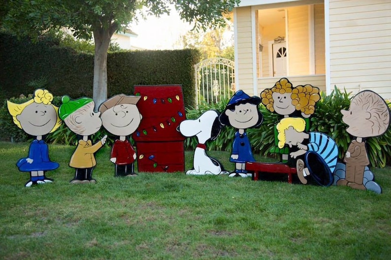 May include: A set of Peanuts characters made of wood and painted in bright colors. The characters include Charlie Brown, Lucy, Linus, Snoopy, and Sally. The characters are standing in a grassy lawn in front of a red and white Christmas tree.