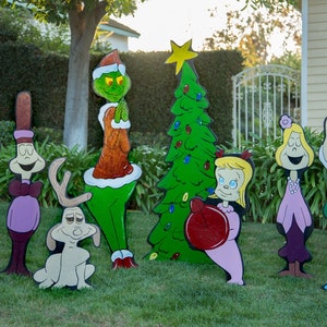 May include: A set of colorful, cartoon-style yard decorations featuring characters from the Grinch and How the Grinch Stole Christmas. The characters include the Grinch, Cindy Lou Who, Max the Dog, and the Whos. The decorations are made of wood and painted in bright colors.